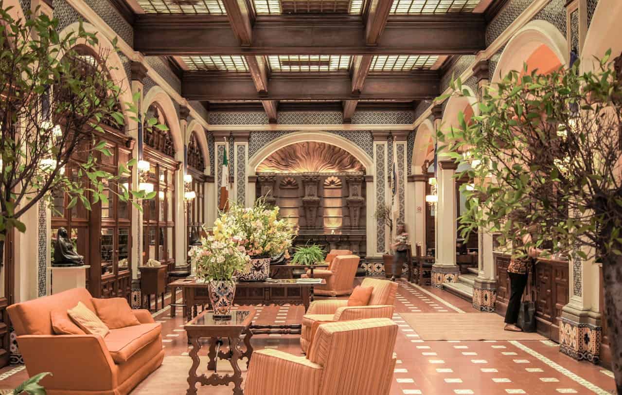 A lavish, high-ceilinged lounge with patterned tile floors, a mix of sofas and armchairs, large potted plants, and decorative ceiling beams, illuminated by ornate light fixtures.