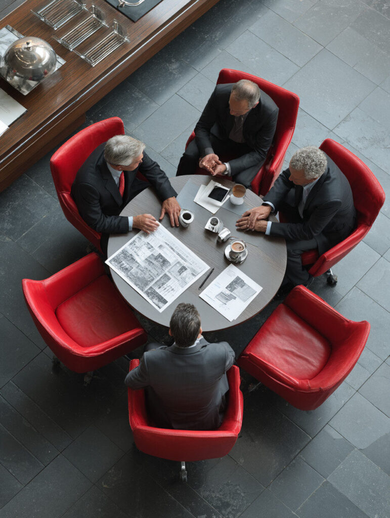 Four men in suits sit around a round table with newspapers, coffee cups, and documents in a modern setting with red chairs, viewed from above.