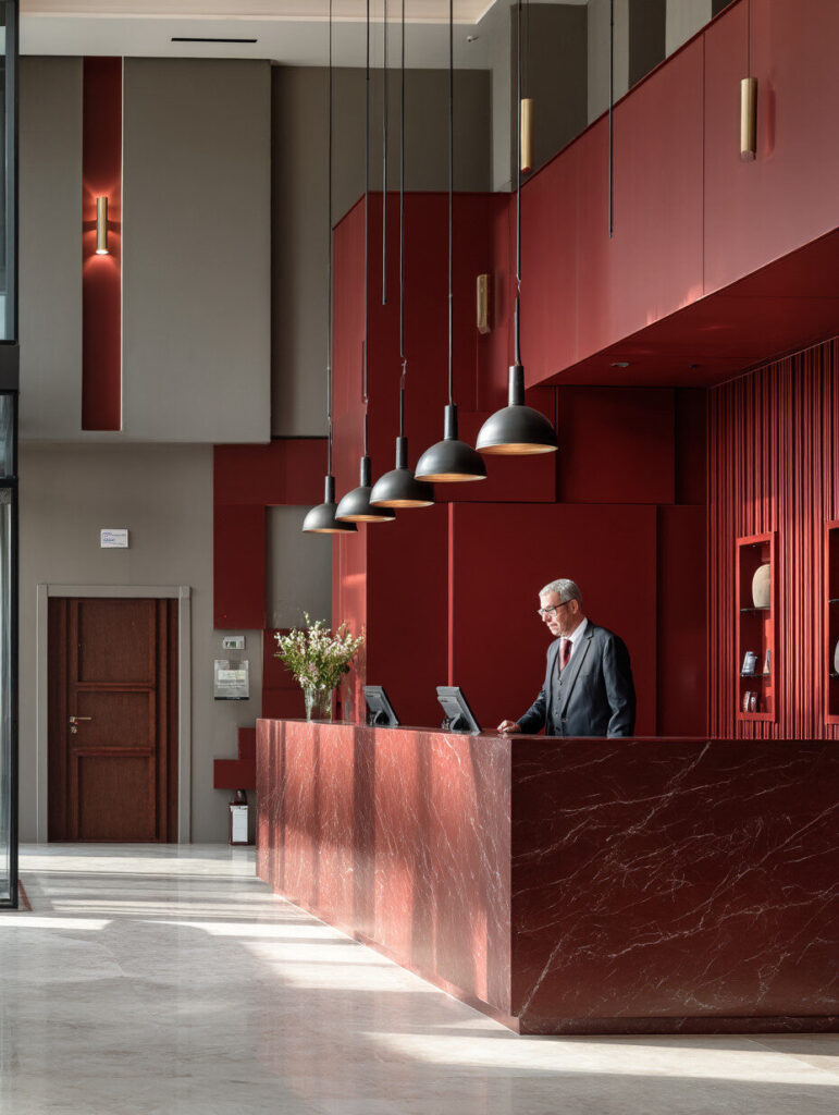 A man in a suit stands behind a modern red marble reception desk in a hotel lobby with pendant lights, tall ceilings, and minimalist decor.